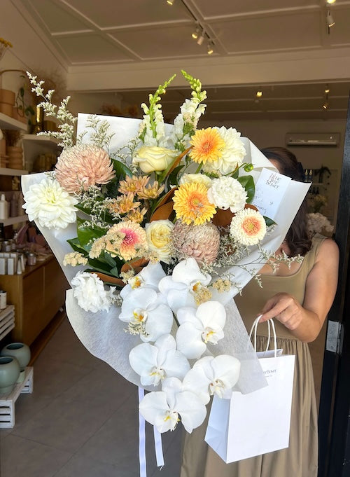 Bouquet of muted, cream, buttery, nude fresh flowers wrapped in white paper, ready for delivery, with a white gift bag held by a person in a store setting.