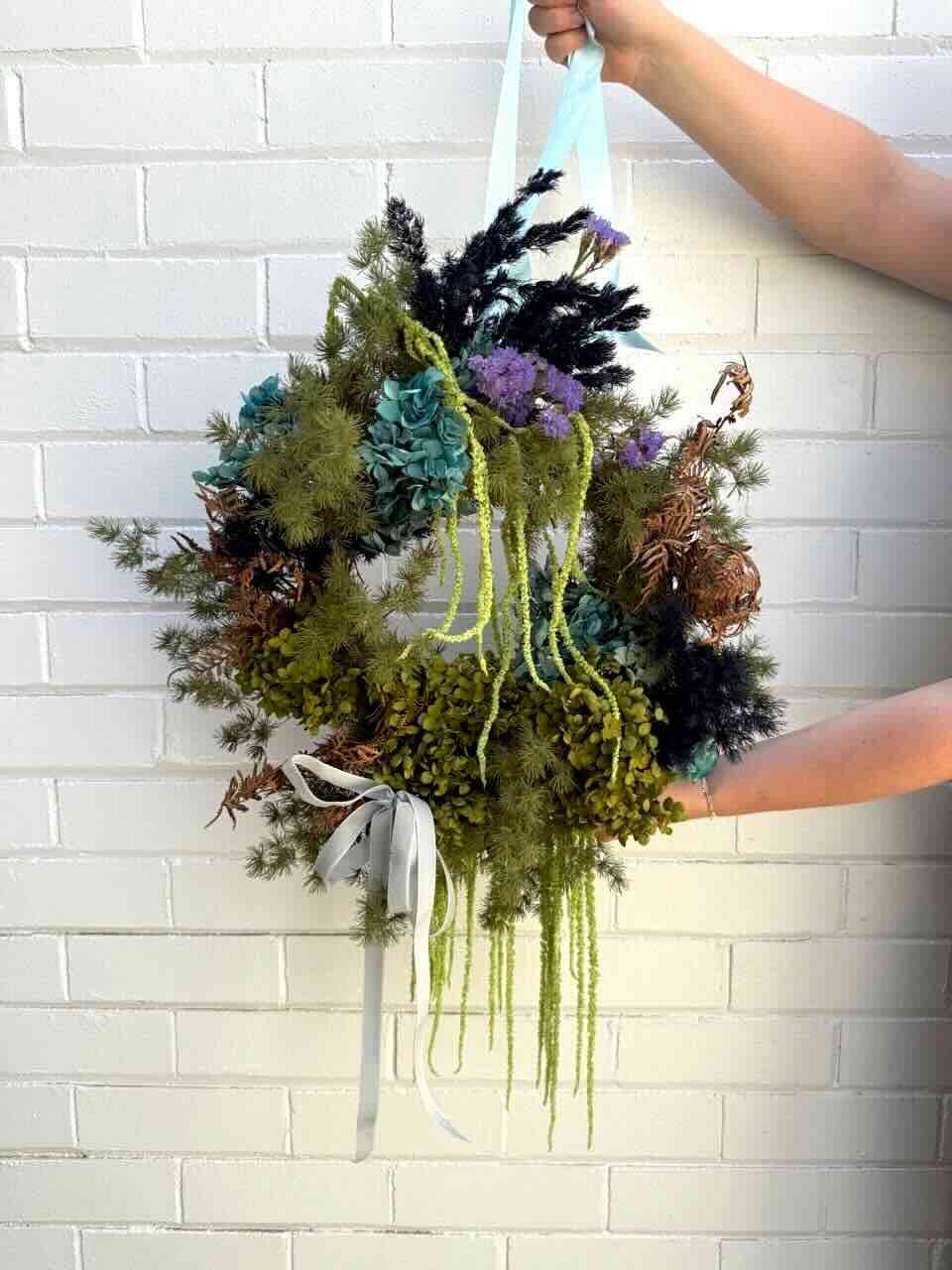A beautifully crafted round Christmas wreath featuring a mix of fresh and dried flowers, greenery, and decorative elements in an asymmetric design, held against a white brick wall.