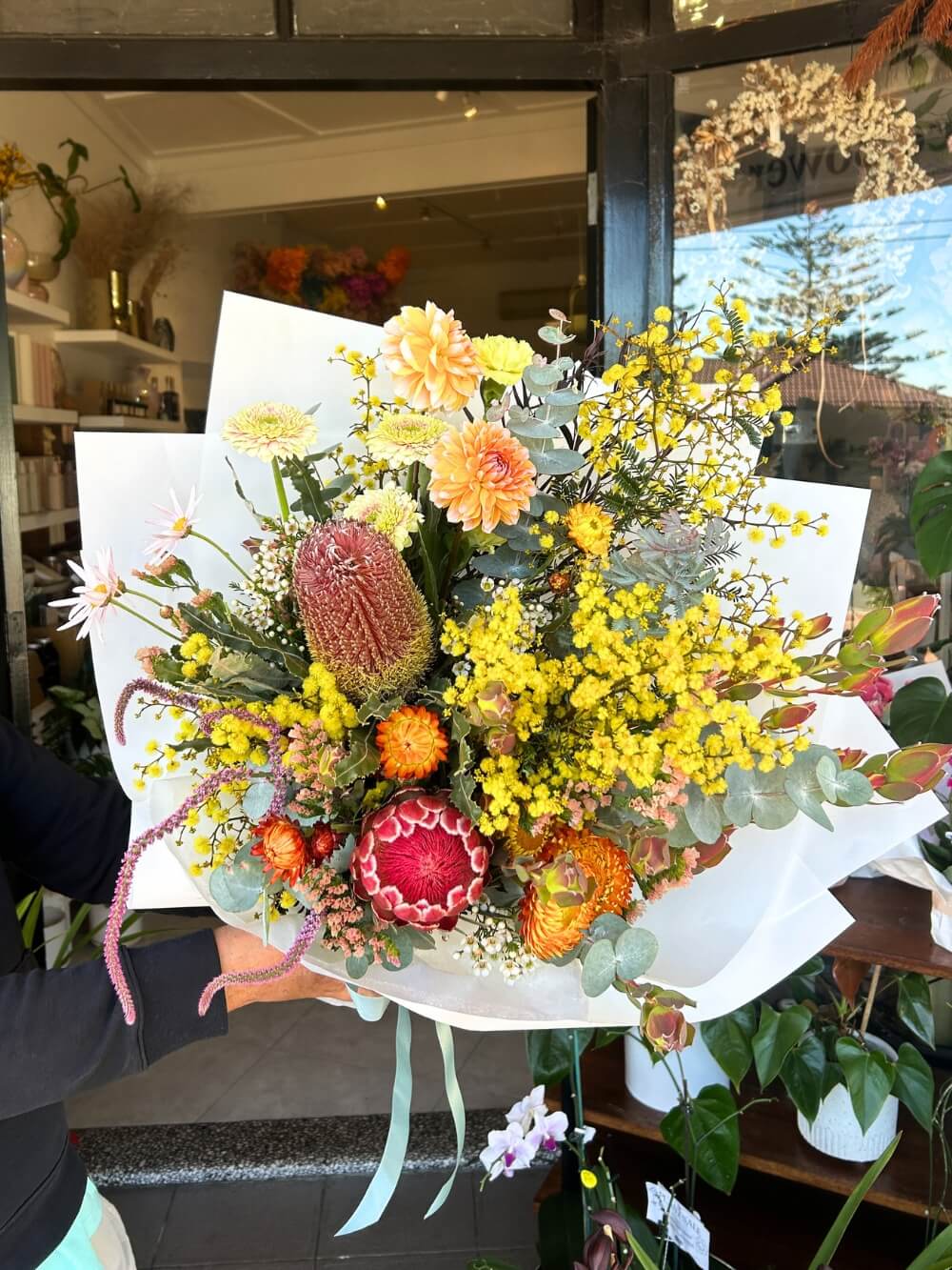 A bouquet of native and rustic flowers including proteas, banksias, and kangaroo paw, wrapped in paper with ribbon.