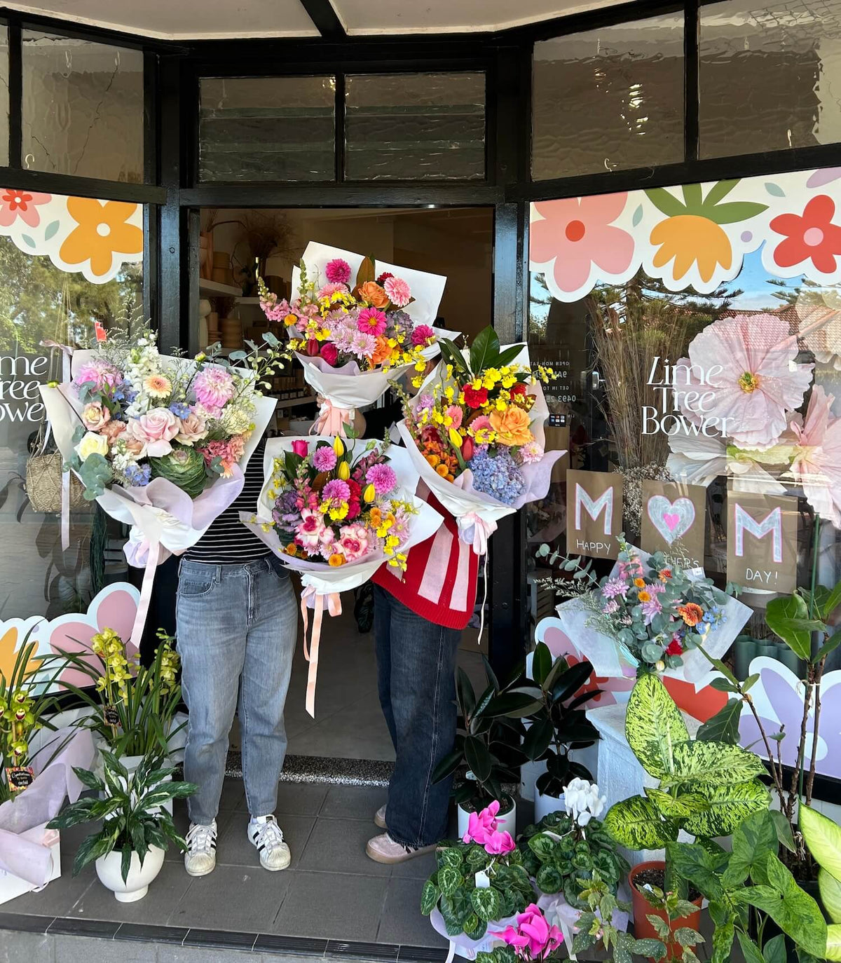 Two people holding flower bouquets in front of a floral shop with decorated windows.