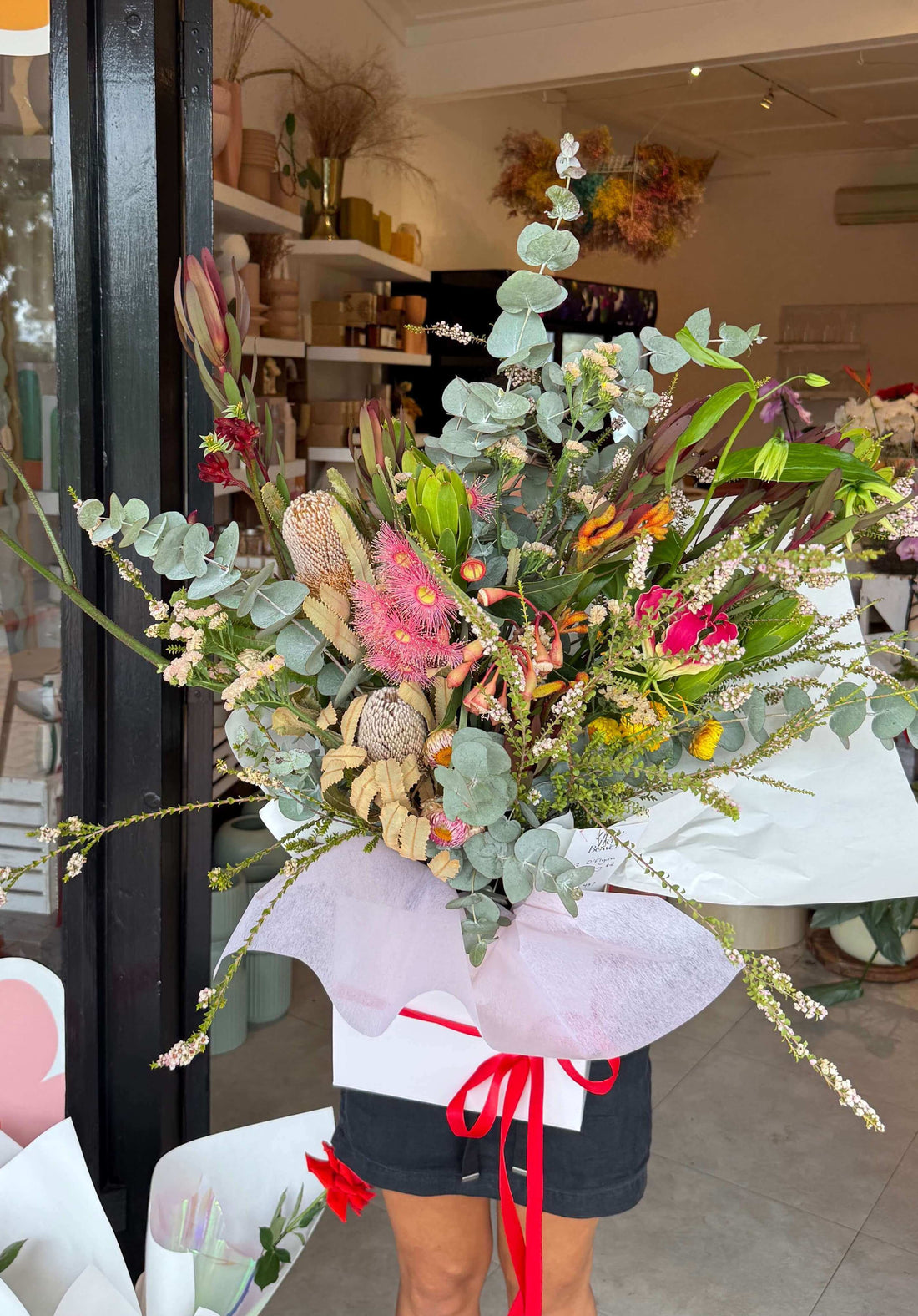 A bouquet of native Australian flowers in various colors, wrapped with a white paper and red ribbon, held by a person in front of a flower shop.