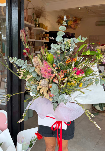A bouquet of native Australian flowers in various colors, wrapped with a white paper and red ribbon, held by a person in front of a flower shop.