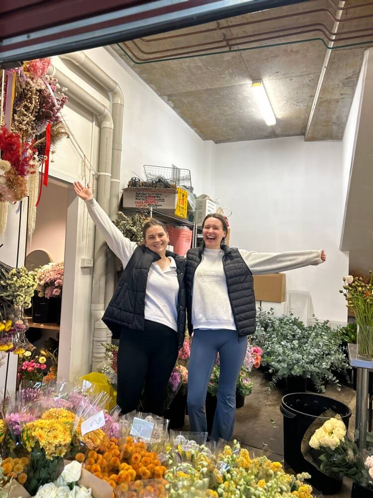 Two female florists standing in a flower shop with arms outstretched, surrounded by flowers for mothers day behind the scenes